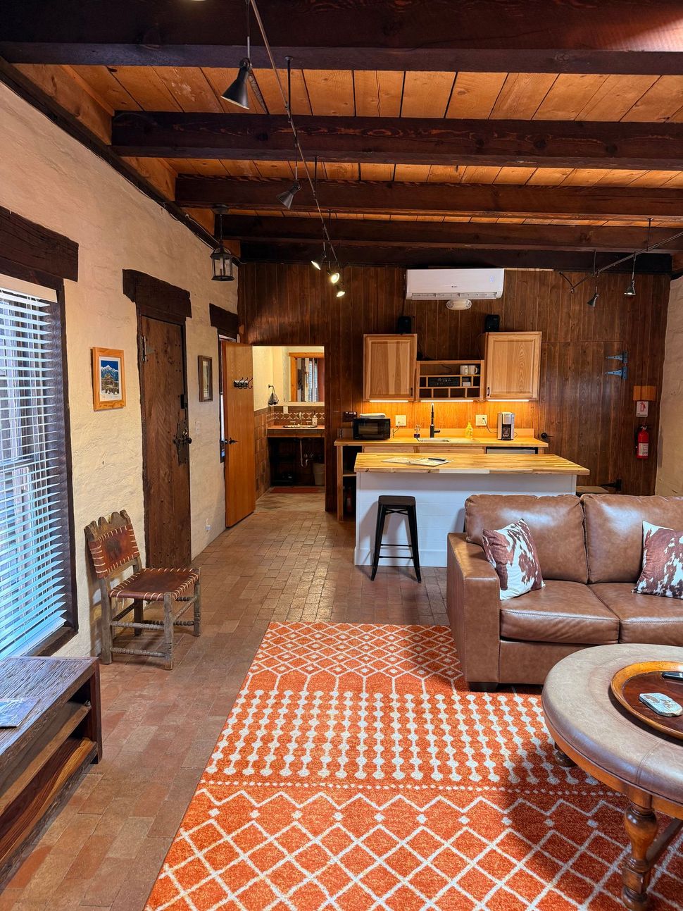Living room with orange rug, leather sofa, kitchen in the background, and wooden beams.