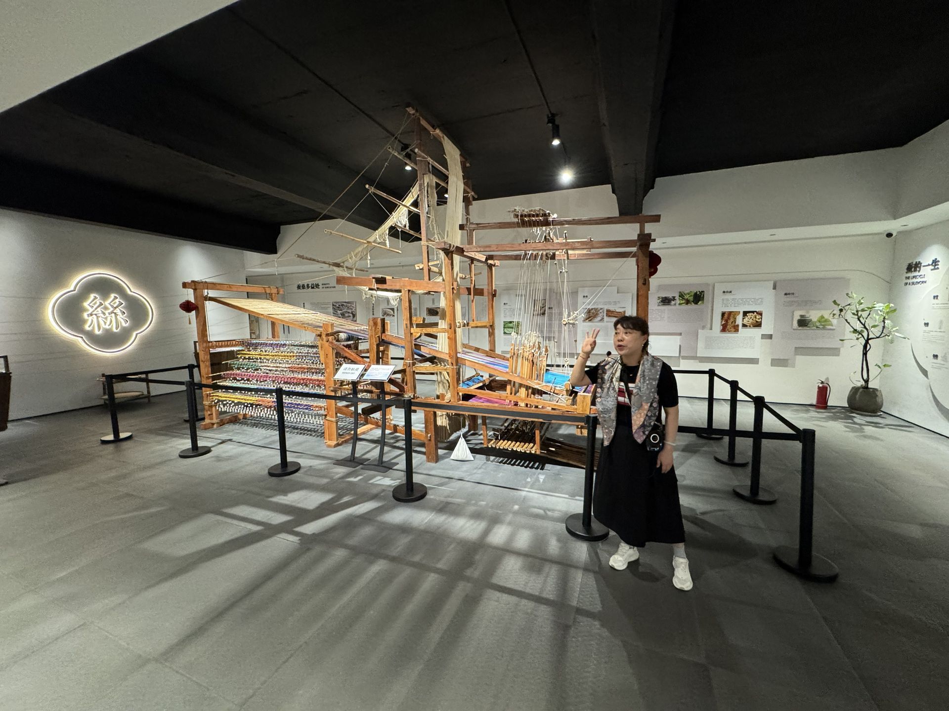 Woman gestures near a large wooden loom in a museum with displays and black barriers.