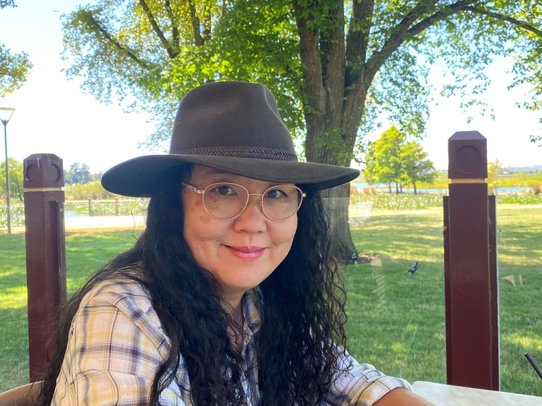 Woman in hat and glasses smiles outdoors at a table, trees in the background.