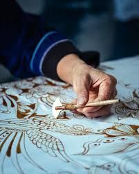 Person's hand using a tool to apply wax to a white batik fabric with a bird design.