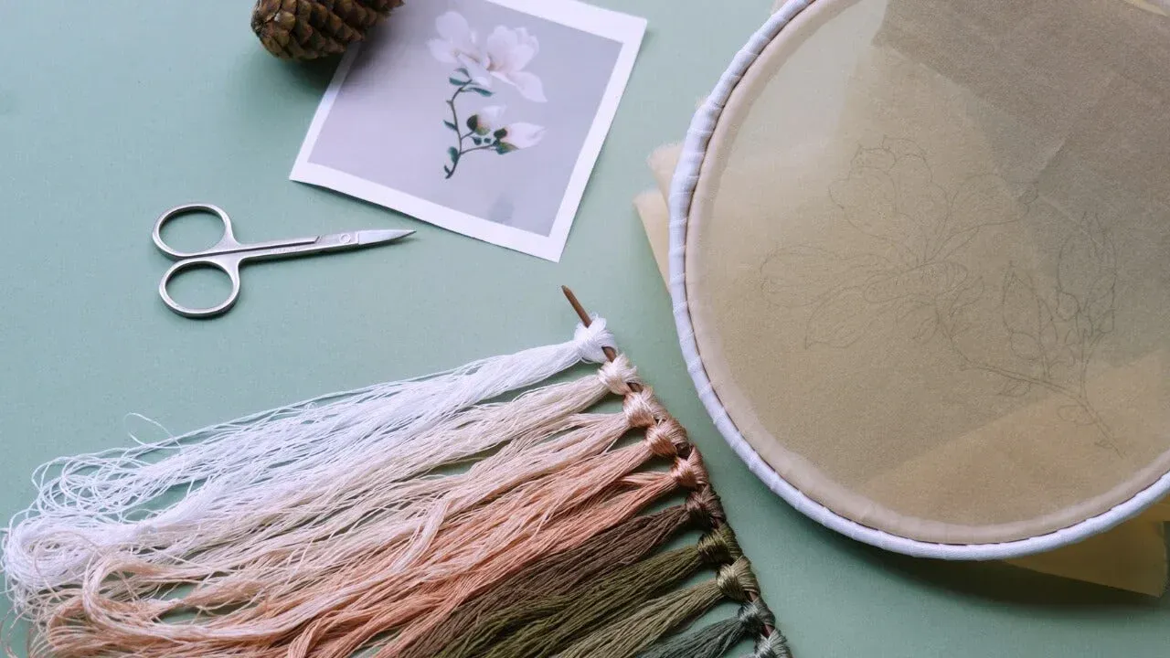 Embroidery supplies: hoop, thread, scissors, and floral design on a pastel green surface.