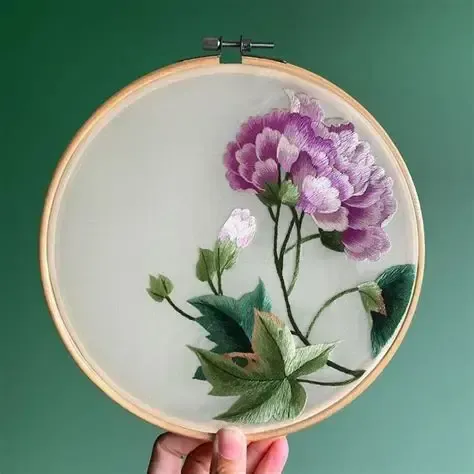 Embroidery hoop with purple flower, green leaves, and buds on translucent fabric. Held against a green background.
