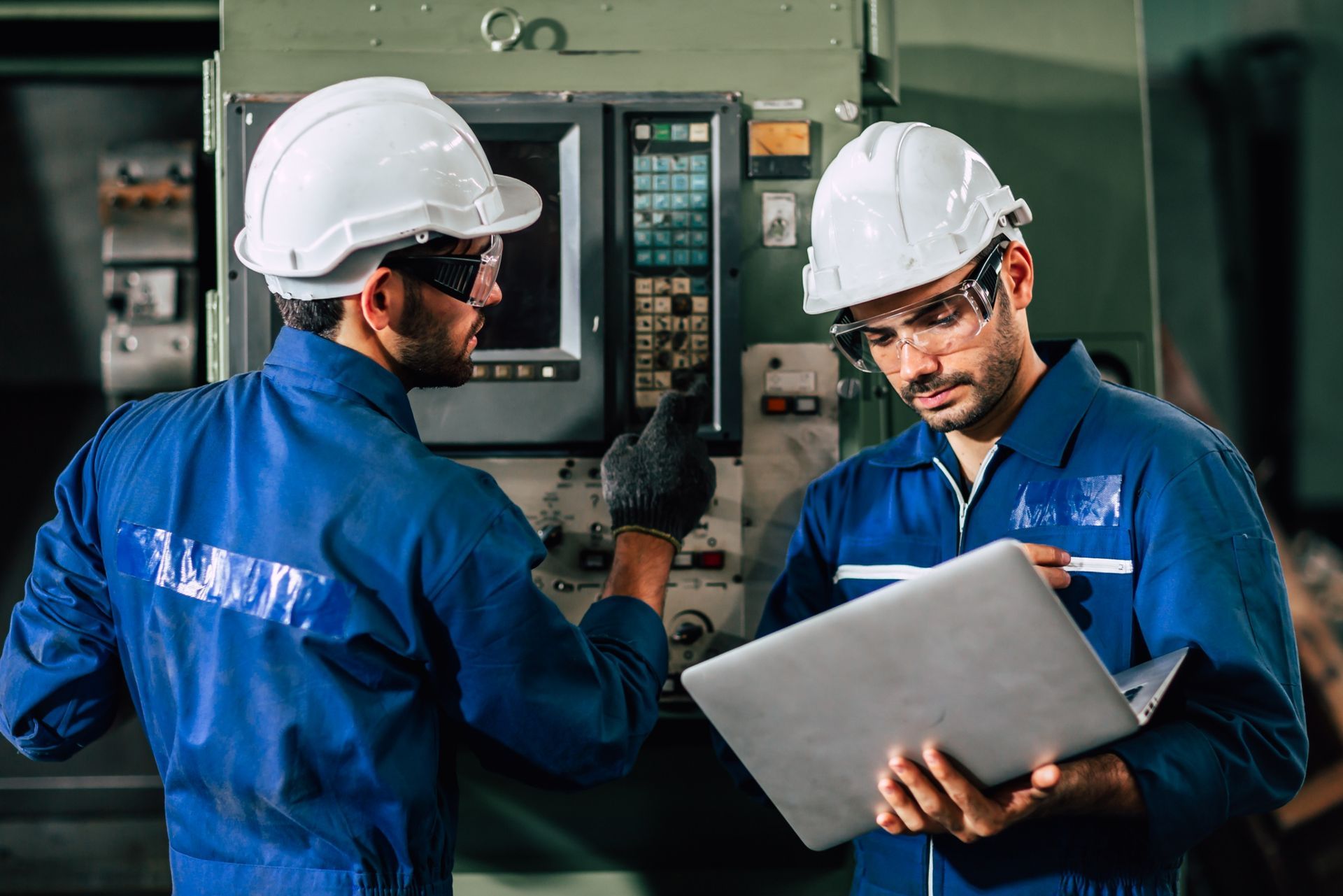 Two workers in blue uniforms and hard hats operate industrial machinery while checking data on a laptop.