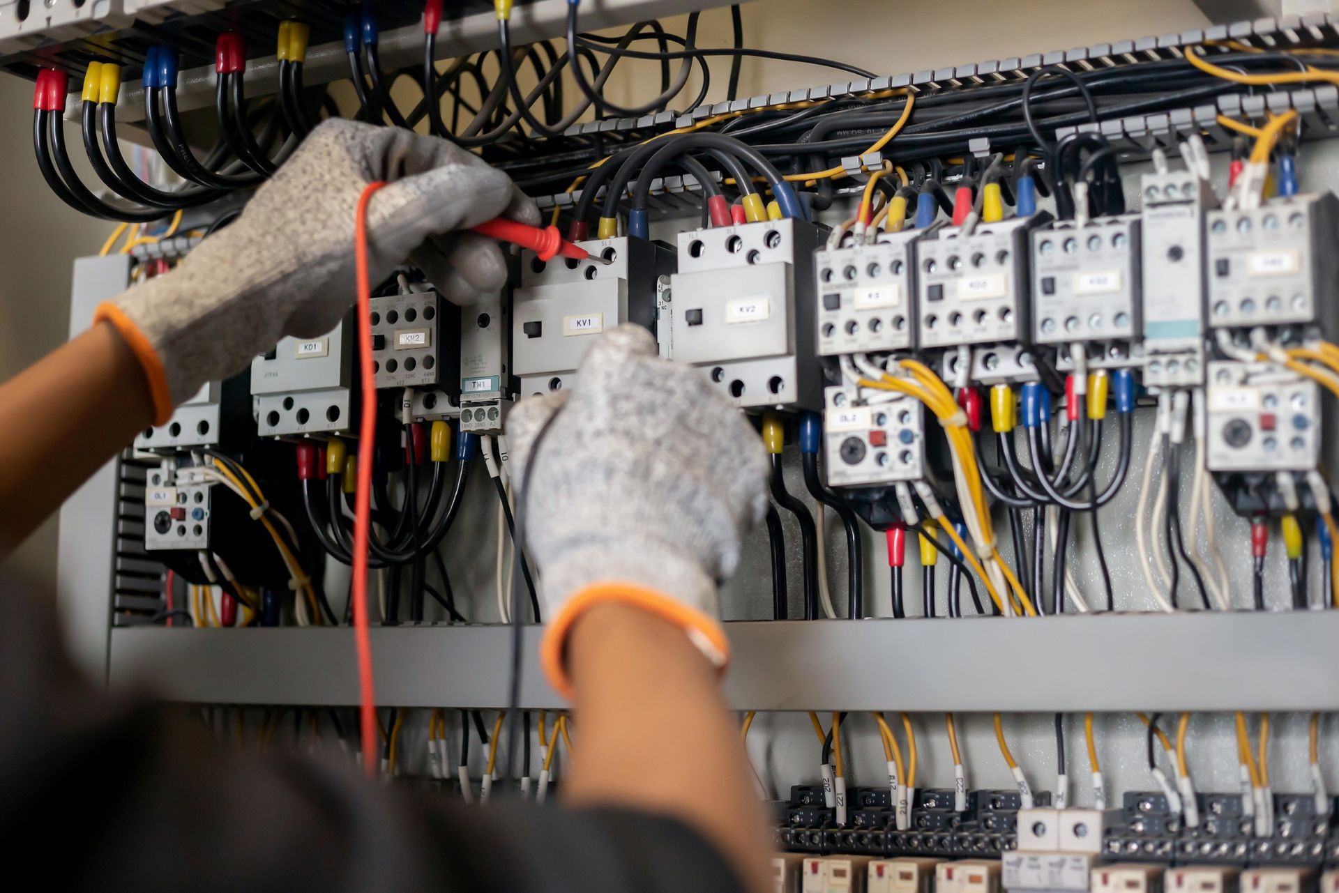 A technician wearing work gloves uses a multimeter probe to test electrical connections on a control panel.