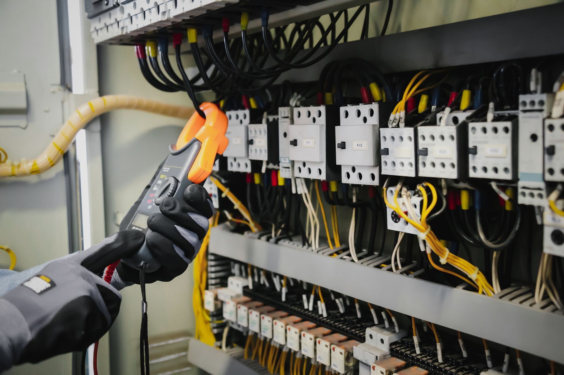 A person wearing gloves uses a clamp meter to measure current on a wire inside an electrical control panel.