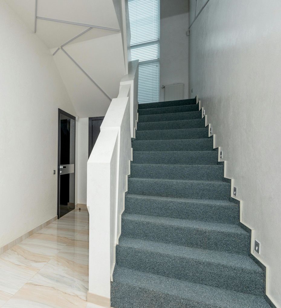 A staircase with blue-gray carpet leads upwards. White walls and a railing border the stairs. A door and window are visible.