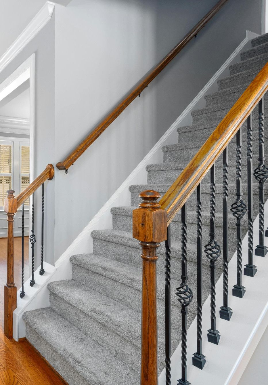 Staircase with gray carpet, wooden handrails, and black wrought iron spindles.