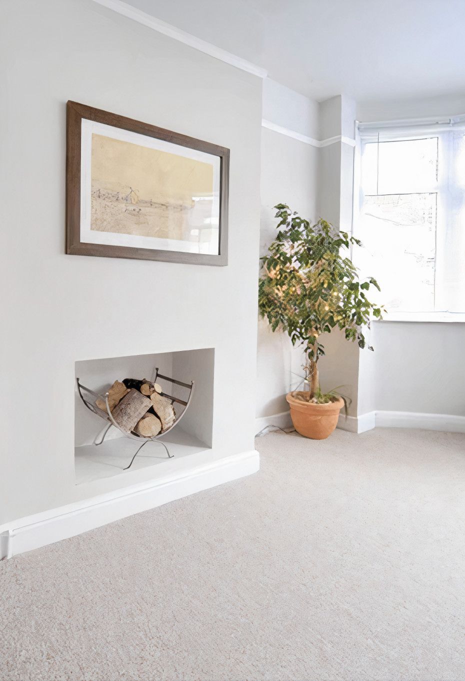 A room with a recessed fireplace holding logs, a framed picture above, and a potted tree near a window. Soft gray walls and carpet.