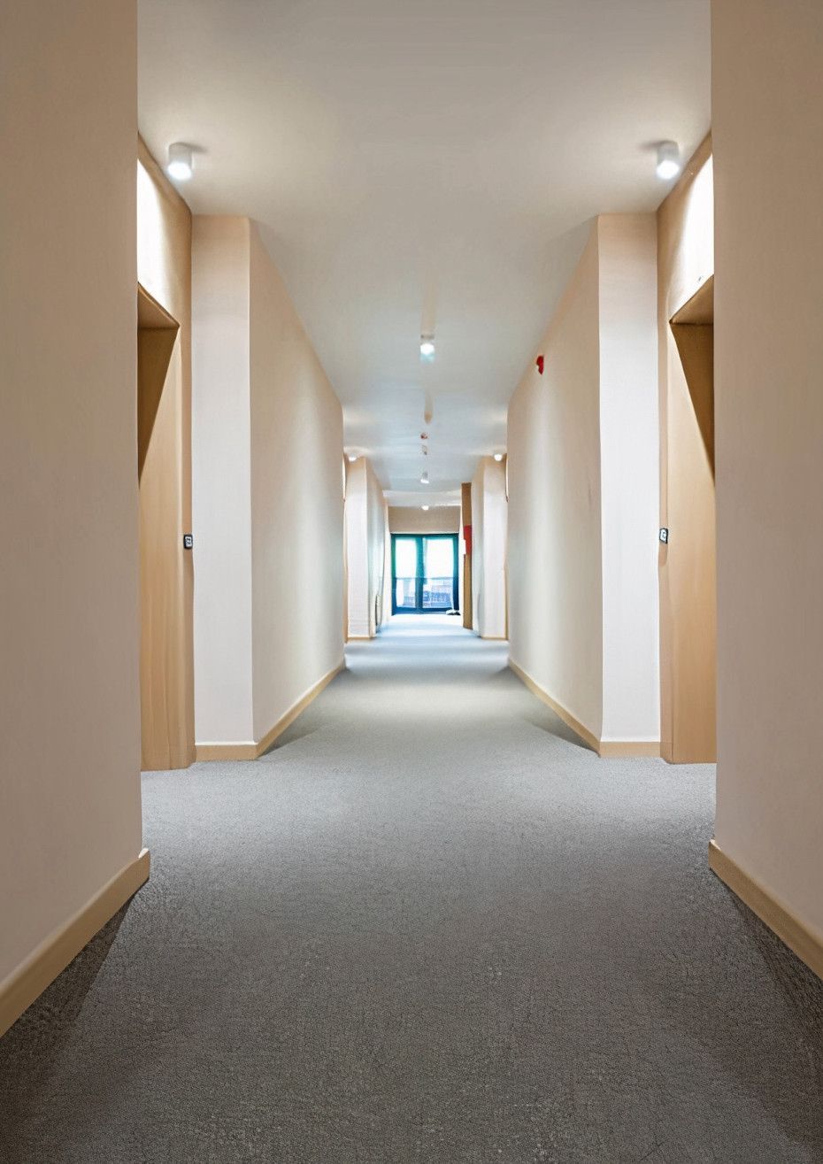 Long, empty hallway with light walls, wooden doors, and gray carpet, leading towards an open doorway at the end.
