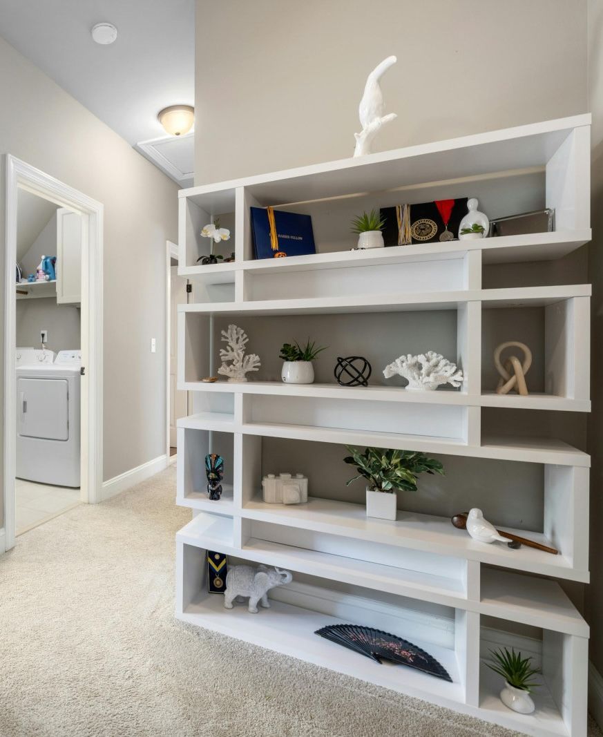 White modern shelving unit in a hallway, filled with decorative objects and plants. The hallway has light beige walls and carpet.