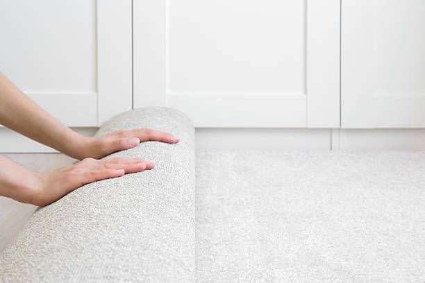 Hands unrolling a light-colored carpet on a white floor, near white cabinets.