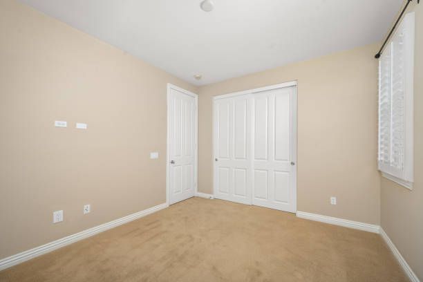 Empty beige bedroom with carpet, two white doors, and a small window with white shutters.