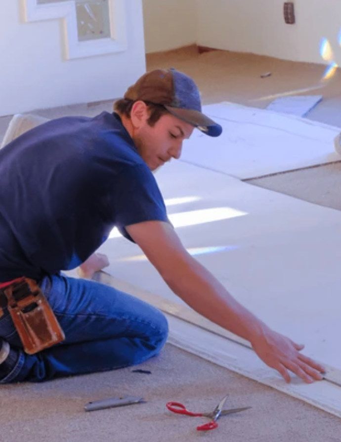 A man in jeans and a blue shirt kneels, measuring flooring. He wears a hat and uses scissors and a utility knife on the light-colored material.