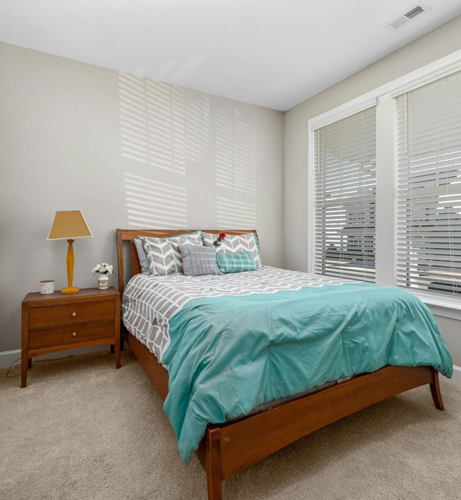 A bedroom with a bed , nightstand , lamp and two windows.