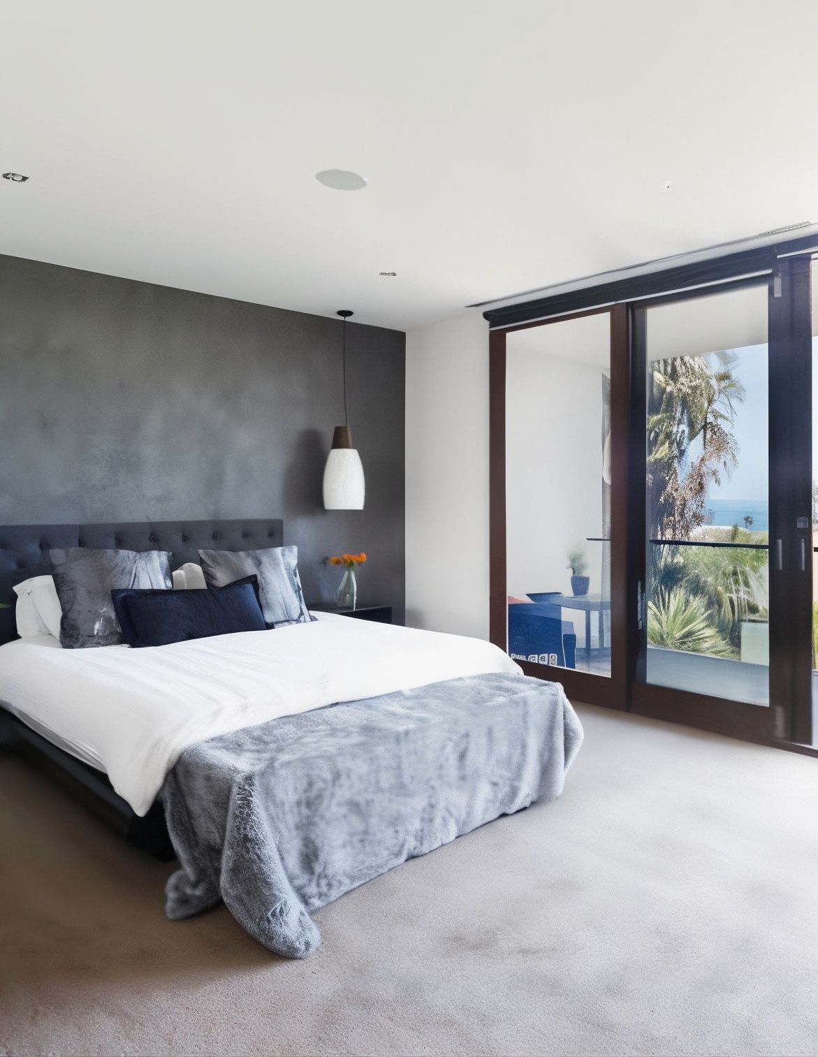 Modern bedroom with a dark gray accent wall behind a bed with gray and white bedding. A large sliding glass door leads to a balcony with trees.