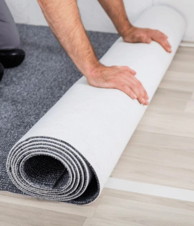 Person rolling out a gray carpet onto a light-colored wooden floor. Hands are visible, and the carpet roll is white.
