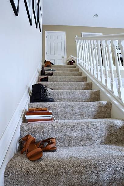 Staircase with shoes and books on the steps. Tan carpeted stairs, white banister, shoes in various colors.