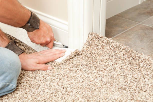 Person trimming carpet with a utility knife near a white wall and tan tile floor.
