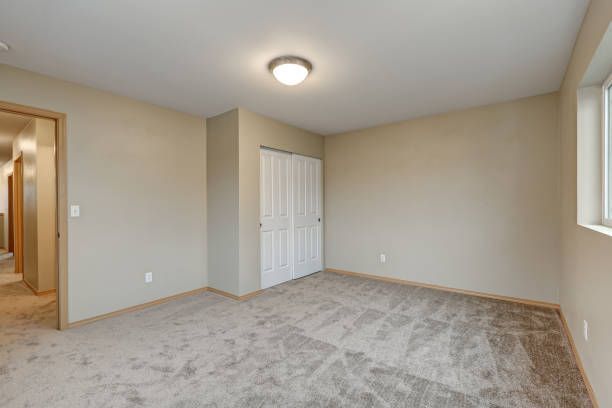 Empty bedroom with beige walls, gray carpet, white closet doors, and a small window.