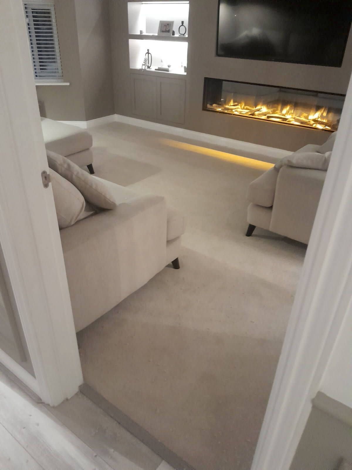 Beige carpeted living room with sectional sofa and fireplace. Built-in shelving and TV on the wall.