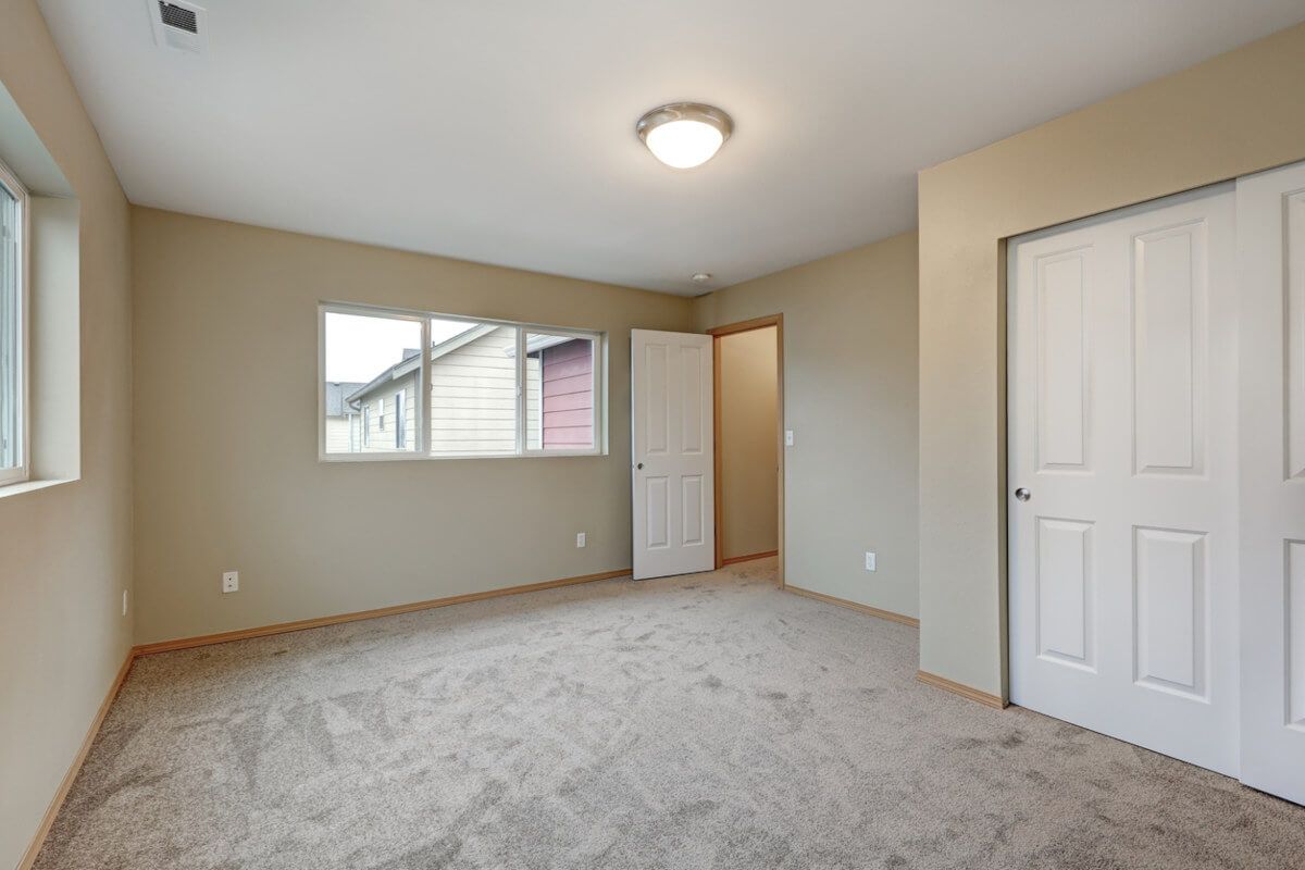 Empty bedroom with beige walls, carpet, windows, and white doors.