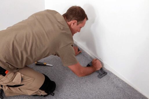 Man installing gray carpet against a white wall, using a tool to secure it.