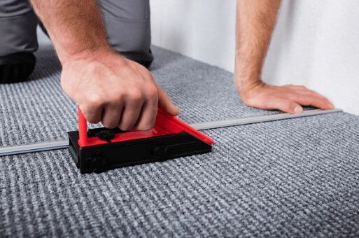 Person using a tool to seam carpet, kneeling on the floor. Red and black tool with grey carpet.