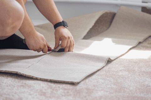 Person kneeling, cutting a carpet with a utility knife. Beige carpet in a bright room.