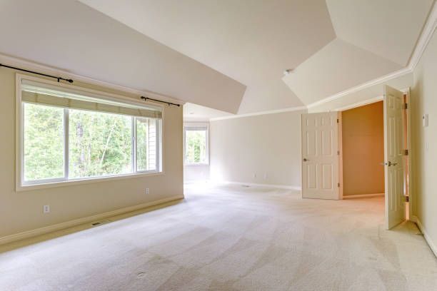 Empty, spacious bedroom with off-white walls, carpet, and a large window overlooking trees.