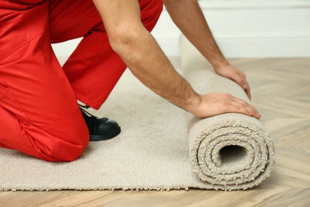 Person in red overalls rolling out beige carpet on a light wood floor.