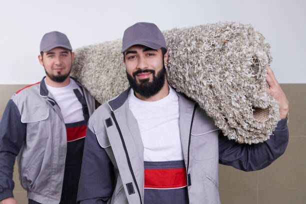 Two men in work uniforms carrying a rolled-up carpet; one has it on his shoulder.