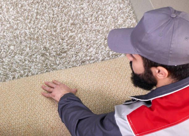 Man in cap examining a carpet, comparing texture, likely assessing for cleaning or installation.