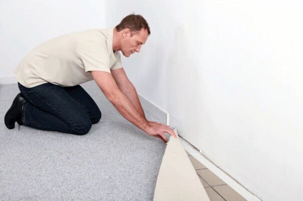 Man kneels, installing baseboard against a white wall. Carpet floor, light-colored baseboard.