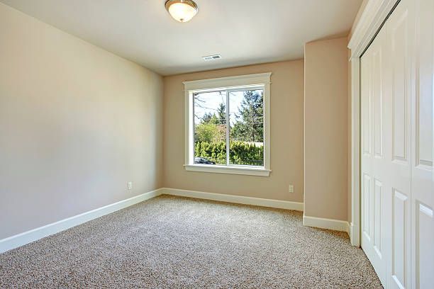 Empty bedroom with beige walls, carpet, and white trim, window, and closet doors.