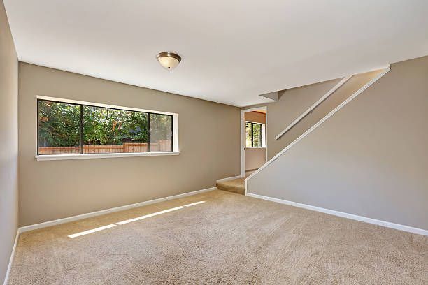Empty room with tan carpet, beige walls, a window, and a staircase. Sunlight streams in.