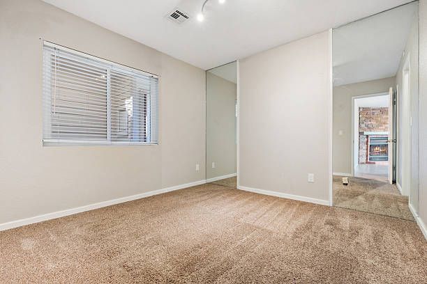 Empty bedroom with beige carpet, blinds, and a hallway visible.