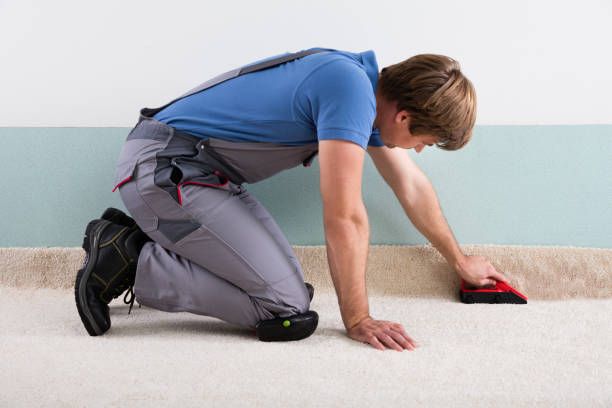 A man kneels, smoothing beige carpet with a red tool against a two-tone wall.