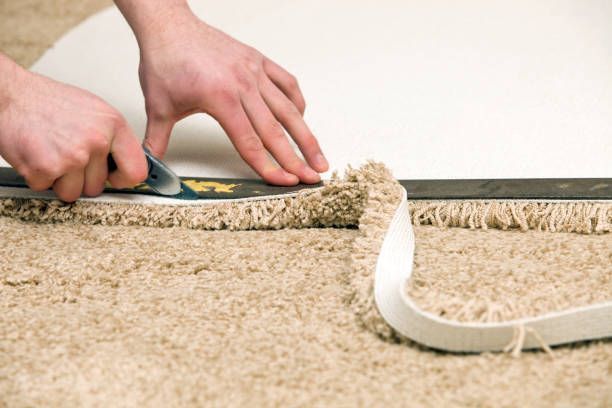 Hands using a cutter to trim carpet along a curved edge, guided by a straight edge.