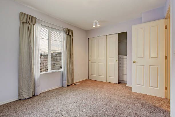 Empty bedroom with beige carpet, closed closet doors, window with curtains, and a closed door.
