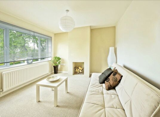 Living room with white sofa, small table, and fireplace. Large window with blinds.