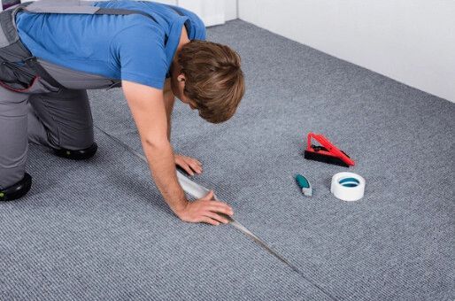 Man kneeling, cutting carpet seam with a knife. Tools and tape beside him, indoors.