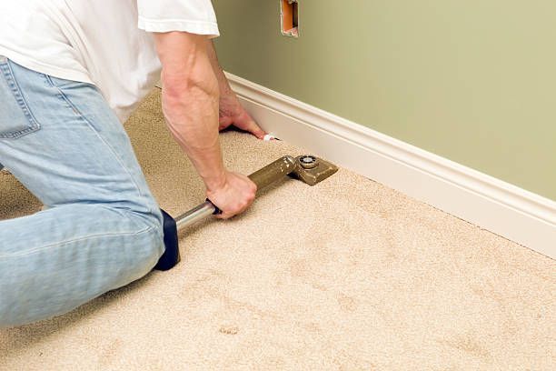 Person kneeling, installing carpet near a wall, using a knee kicker tool.