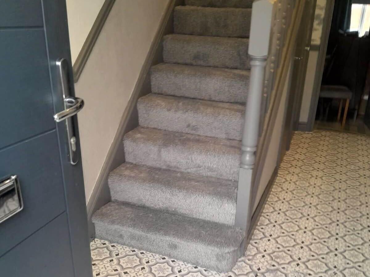 Gray carpeted staircase with gray banister next to a patterned floor and a blue door.