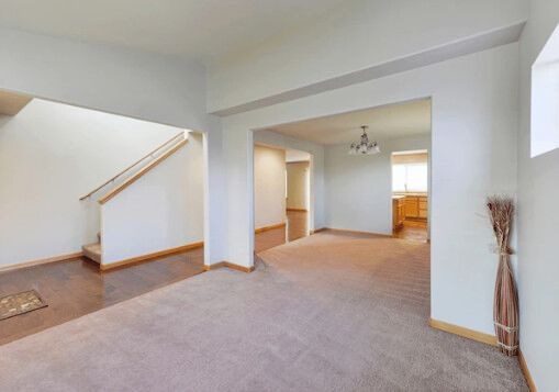 Empty living room with carpet and beige walls; stairs to the left, kitchen through an archway.