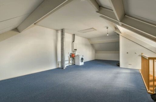 Empty attic room with blue carpet, white walls, and wooden beams.