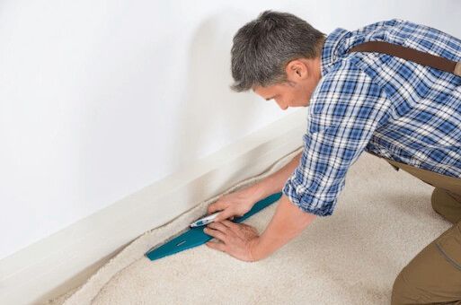 Man kneeling, using a carpet knife to trim carpet along a white wall baseboard.