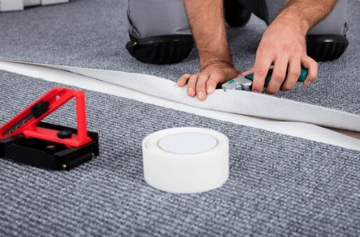 Person cutting carpet with a utility knife, tools and tape on gray carpet floor.