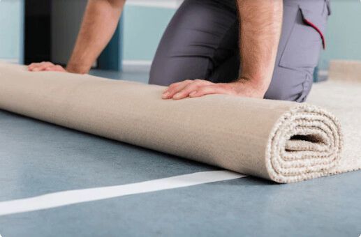Person kneeling, unrolling a beige carpet on a blue floor. White tape marks the edge.