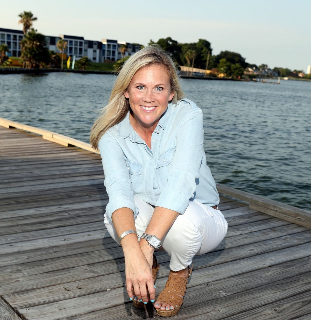 Woman in light blue shirt and white pants crouches on a wooden dock by the water, smiling.