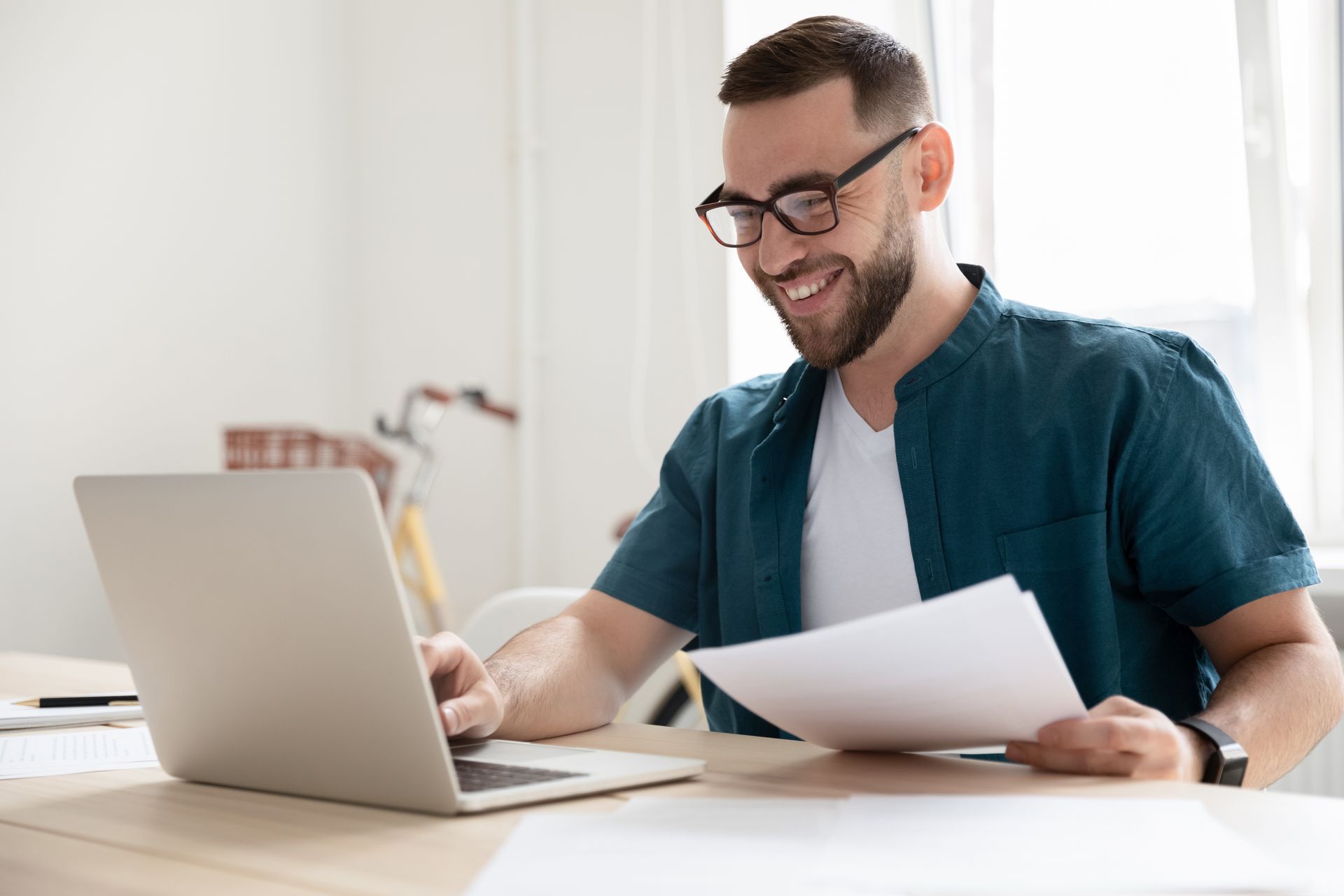 A man is sitting at a desk using a laptop computer and holding a piece of paper.